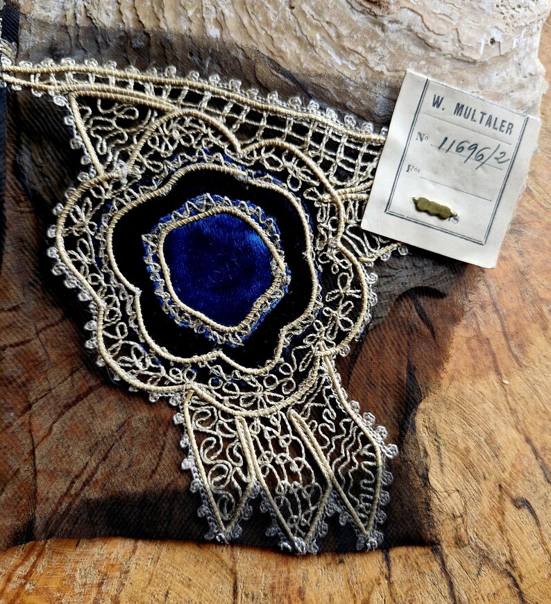 May include: A close-up of a gold and silver lace trim with a blue velvet center. The trim is on a black fabric background. A tag with the text "W. M&Uuml;LTALER No. 11696/2" is visible in the upper right corner of the image.