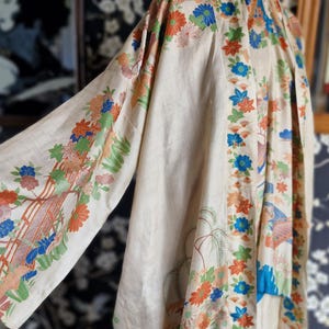 May include: Cream-colored kimono with floral and landscape patterns in orange, blue, and green. The sleeves and edges are adorned with detailed designs. The garment is displayed on a mannequin, showcasing its flowing fabric and intricate artwork.