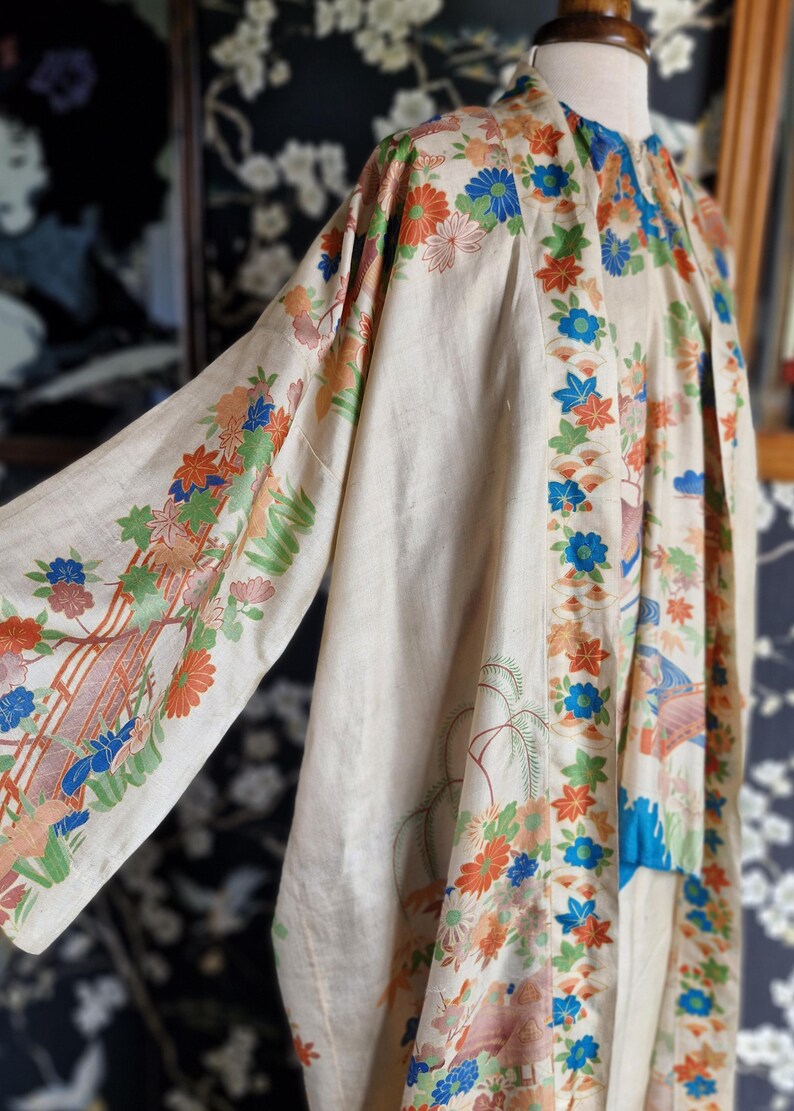 May include: Cream-colored kimono with floral and landscape patterns in orange, blue, and green. The sleeves and edges are adorned with detailed designs. The garment is displayed on a mannequin, showcasing its flowing fabric and intricate artwork.