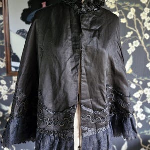 May include: A black satin cape with a lace trim and a ruffled collar. The cape is displayed on a mannequin.