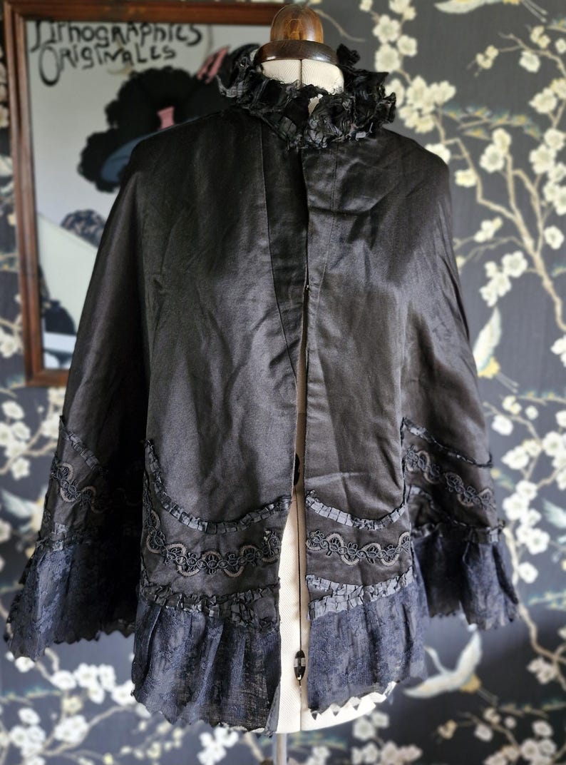May include: A black satin cape with a lace trim and a ruffled collar. The cape is displayed on a mannequin.