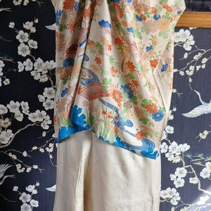 May include: Cream-colored silk ensemble featuring a top and pants. The top has a floral design with orange, blue, and green accents. The pants have a matching floral border. The outfit is displayed on a mannequin.