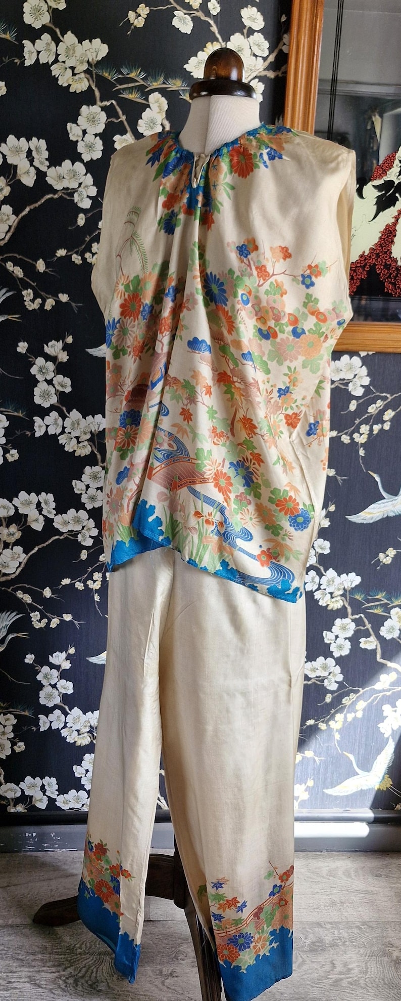 May include: Cream-colored silk ensemble featuring a top and pants. The top has a floral design with orange, blue, and green accents. The pants have a matching floral border. The outfit is displayed on a mannequin.