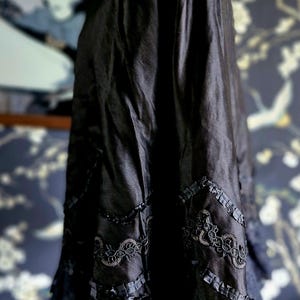 May include: A black silk cape with lace trim and floral embroidery. The cape is draped over a wooden mannequin.