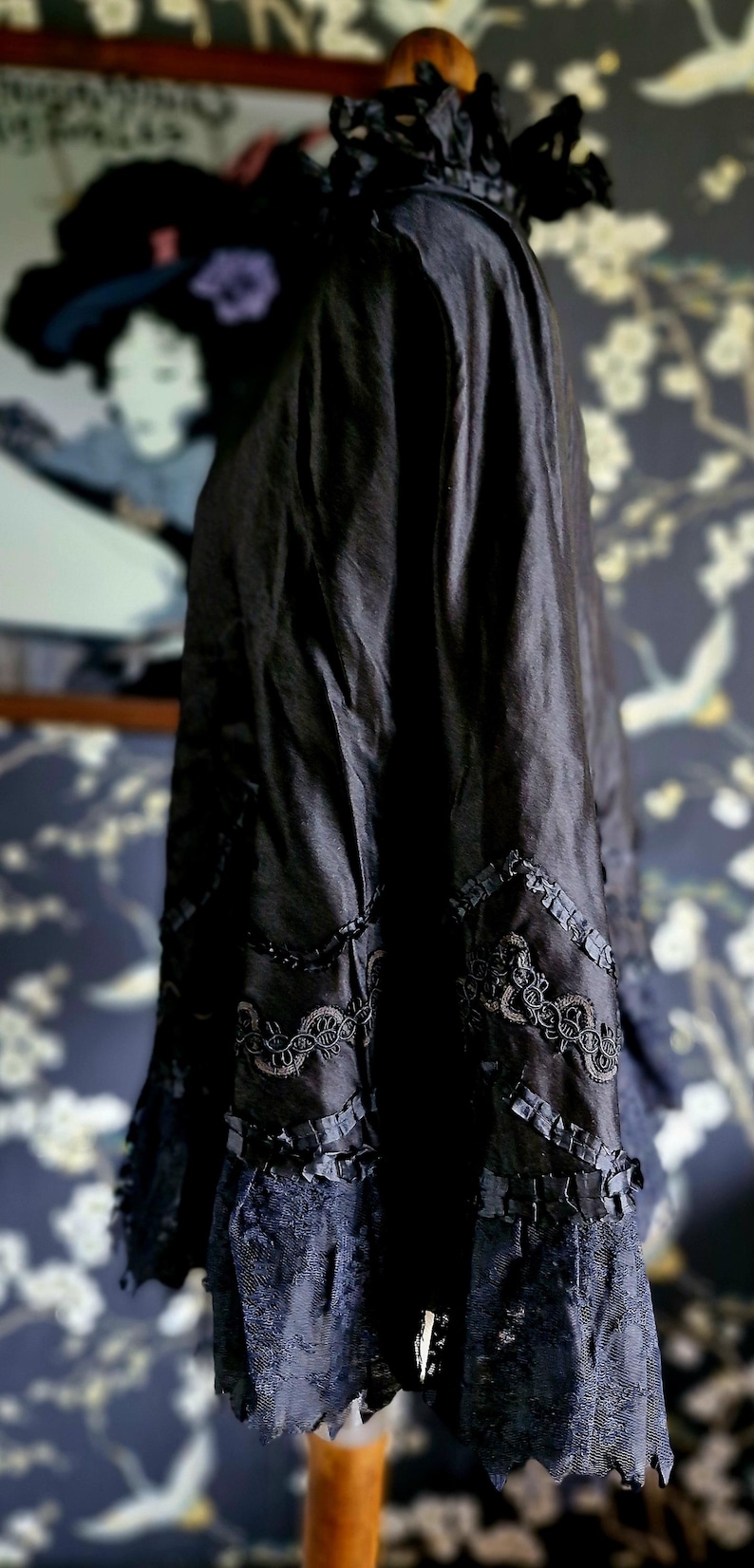 May include: A black silk cape with lace trim and floral embroidery. The cape is draped over a wooden mannequin.