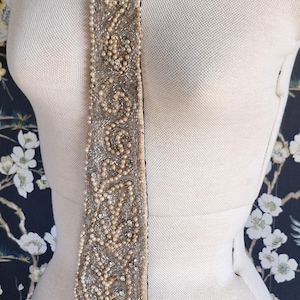 May include: A silver beaded sash with pearl accents and a fringe. The decorative belt is displayed on a mannequin. The sash is embellished with intricate patterns and hangs vertically.