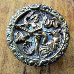 May include: A circular brass button with an ornate design featuring a figure holding a spear and a mythical creature. The button has a green patina and intricate details.