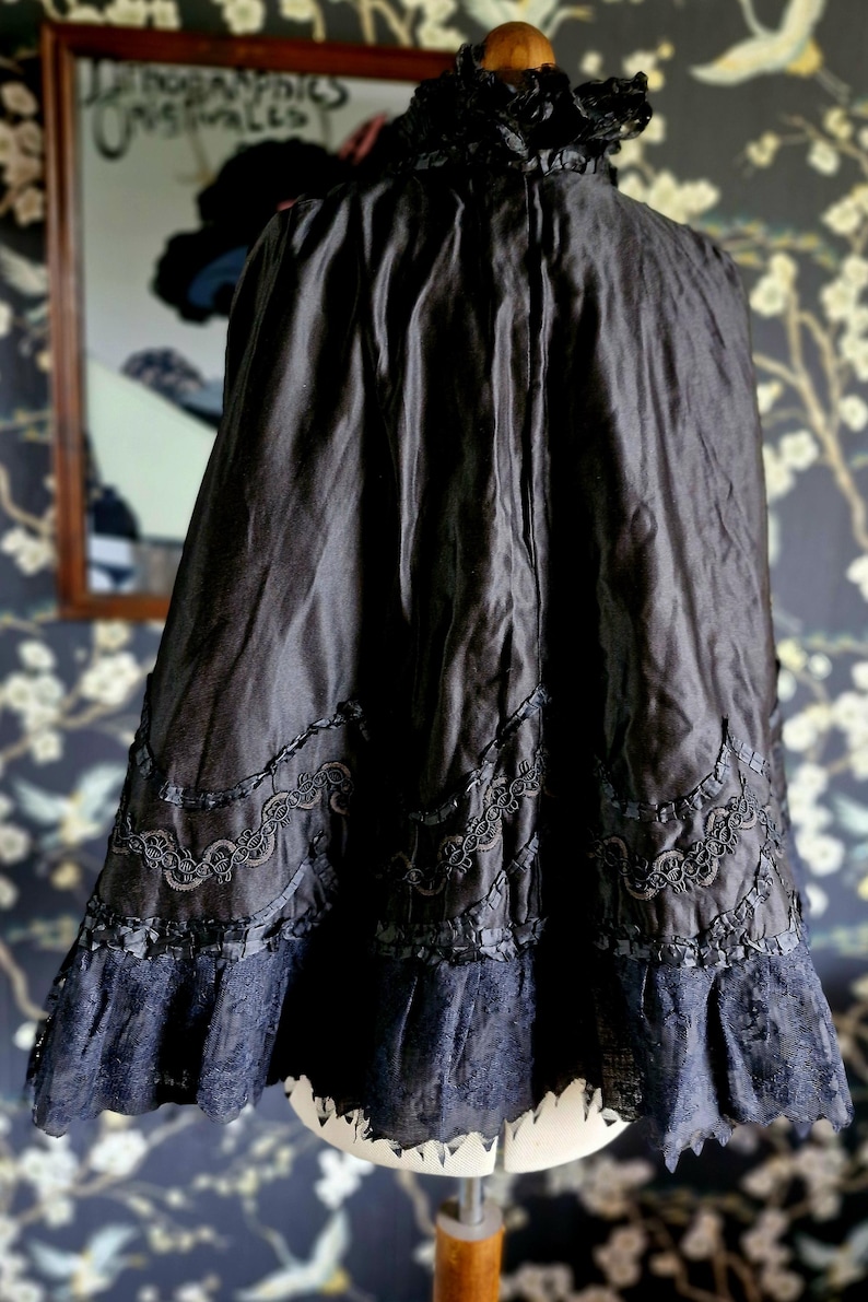 May include: A black satin cape with a high ruffled collar and lace trim. The cape is lined with black fabric and has a scalloped hem.