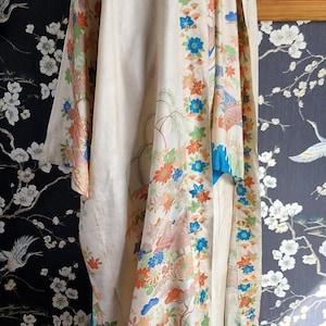 May include: Cream-colored kimono with floral and bird patterns in orange, blue, and green. The kimono has a blue border and is displayed on a mannequin. The background features a floral wallpaper.
