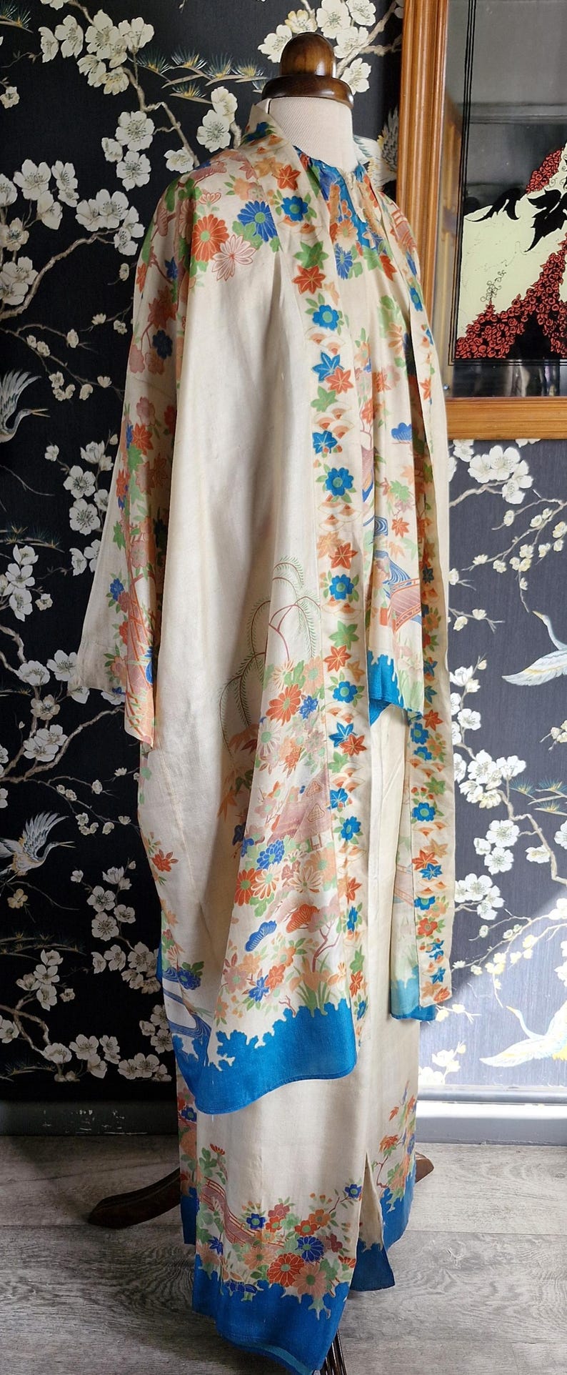 May include: Cream-colored kimono with floral and bird patterns in orange, blue, and green. The kimono has a blue border and is displayed on a mannequin. The background features a floral wallpaper.