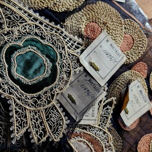 May include: Close-up of several antique fabric trims with gold and silver embroidery. The trims are on a black fabric background. There are three paper tags with the text "W. MULTALER" and numbers.