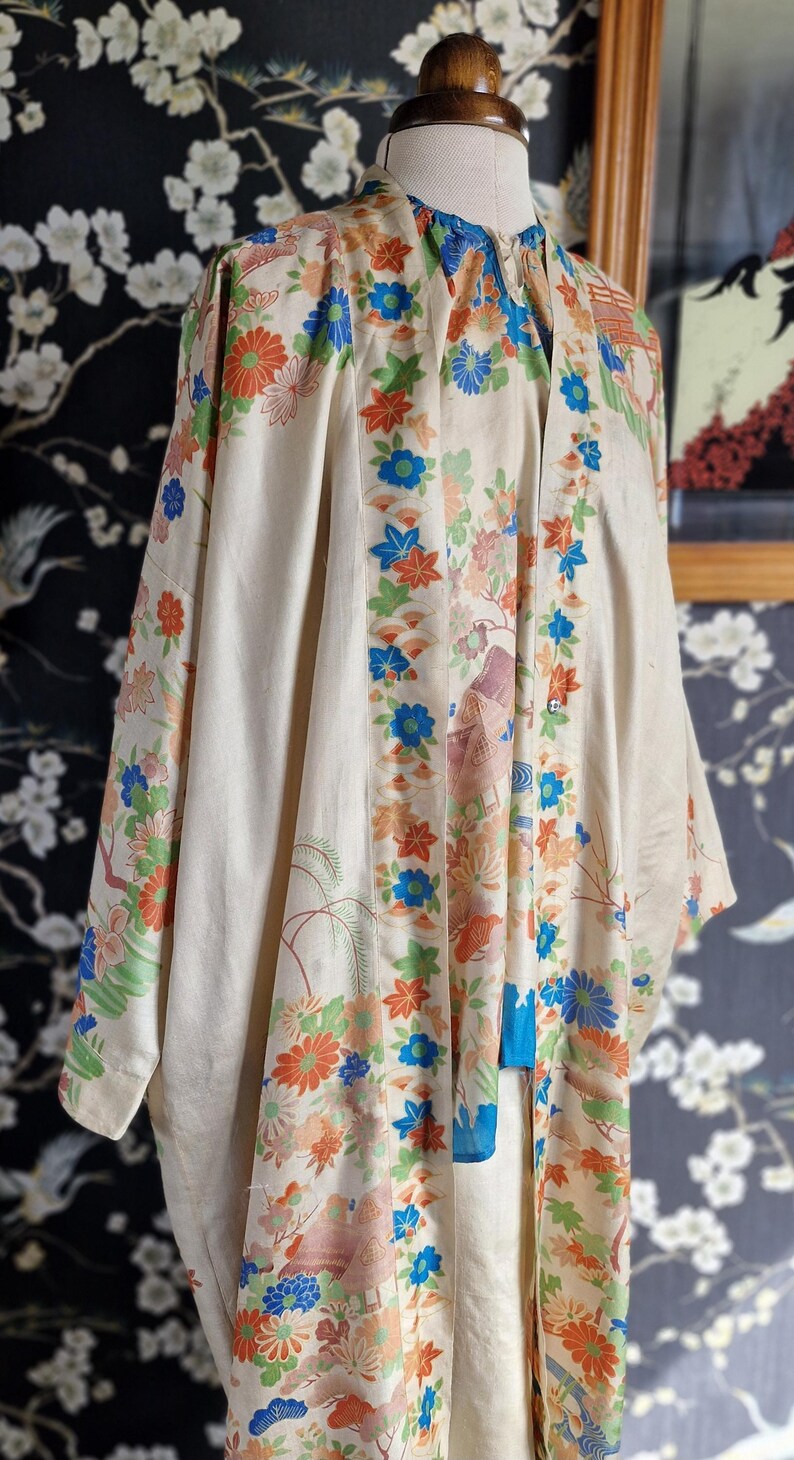 May include: Cream-colored kimono with a floral pattern in orange, blue, and green. The garment features a mandarin collar and long sleeves. The design includes stylized flowers and landscape elements.