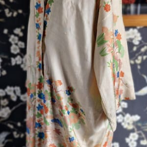 May include: Cream-colored vintage kimono with floral and landscape patterns in orange, blue, and green. The kimono is displayed on a mannequin, showcasing its flowing design and intricate details. The background features a dark floral wallpaper.