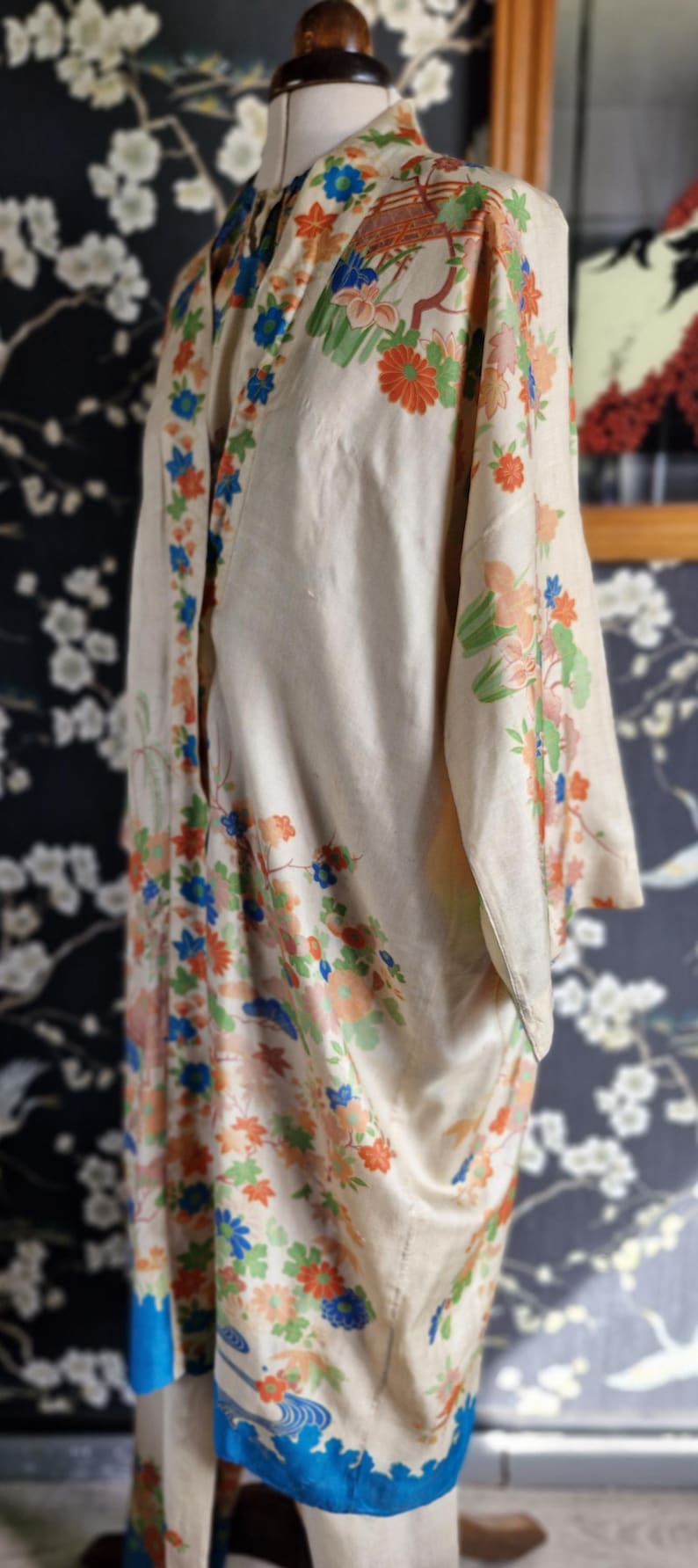 May include: Cream-colored vintage kimono with floral and landscape patterns in orange, blue, and green. The kimono is displayed on a mannequin, showcasing its flowing design and intricate details. The background features a dark floral wallpaper.