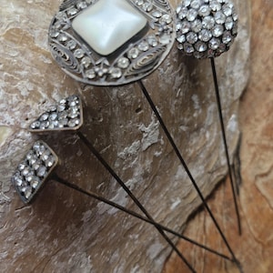 Peut inclure: Un ensemble de quatre épingles à chapeau avec des montures en métal argenté. Une épingle présente un grand cadre circulaire en filigrane avec un cabochon blanc central et des strass, tandis que les autres arborent des motifs géométriques.