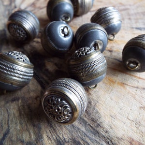 May include: A collection of ten round, silver and gold buttons with intricate designs. The buttons have a dark gray center and are arranged on a wooden surface.