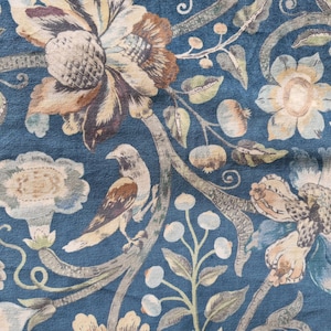 May include: Close-up of a fabric with a floral pattern in shades of blue, brown, and cream. The design features large flowers, leaves, and a bird perched on a branch. The fabric has a textured appearance.