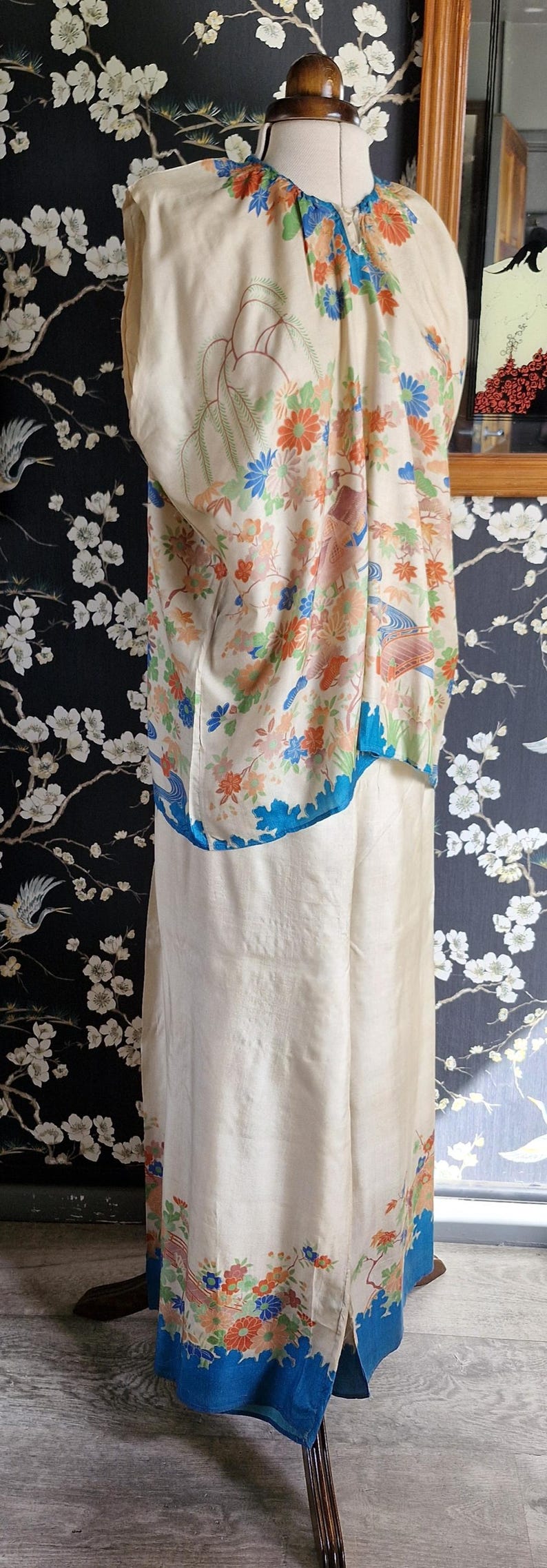 May include: Cream-colored vintage kimono with floral and bird pattern. The kimono features a blue trim and is displayed on a mannequin. The background has a floral design. A unique piece of vintage apparel.