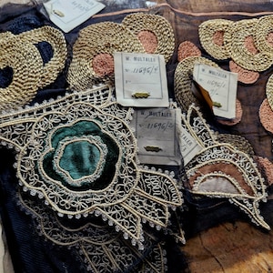 May include: Close-up of several antique lace trims with gold and silver thread embroidery. The trims feature a variety of floral designs, including a green velvet rose.  The trims are labeled with the name "W. Multaler" and a number, "11696/2".
