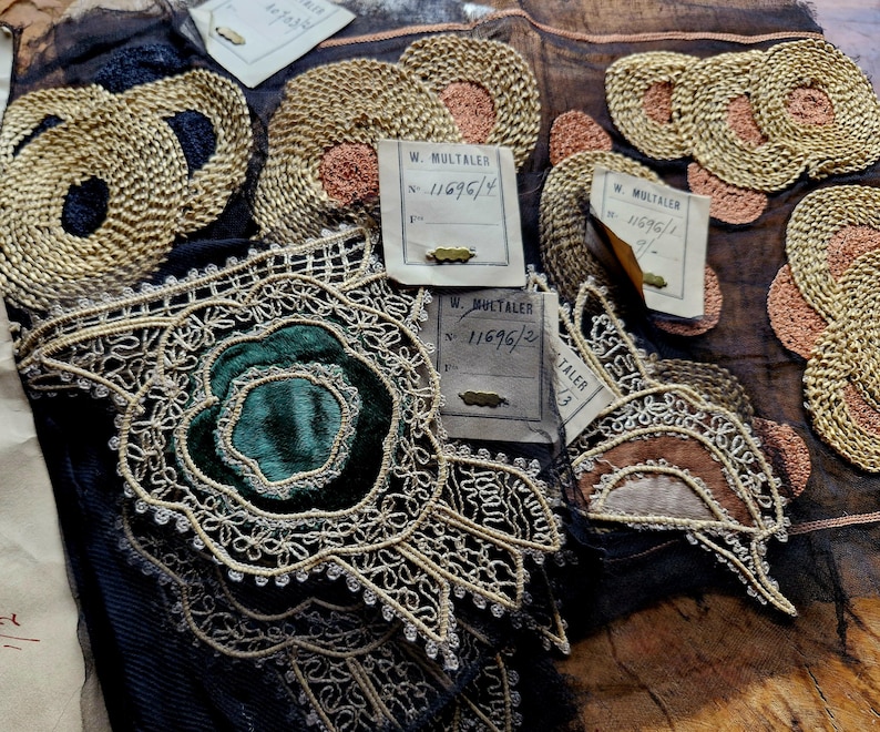 May include: Close-up of several antique lace trims with gold and silver thread embroidery. The trims feature a variety of floral designs, including a green velvet rose.  The trims are labeled with the name "W. Multaler" and a number, "11696/2".