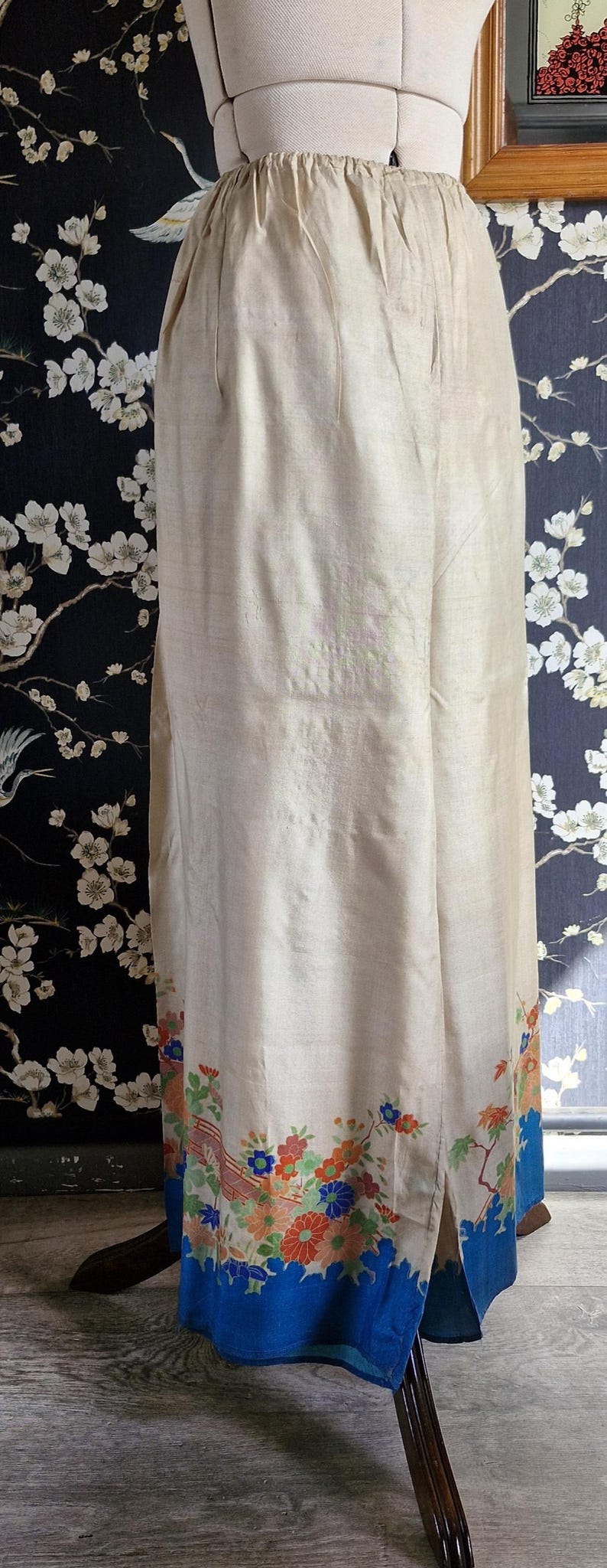 May include: Cream-colored, vintage-style pants with a floral border in shades of blue, orange, and red. The pants are displayed on a mannequin, showcasing the design and fit. The bottom hem is a vibrant blue.