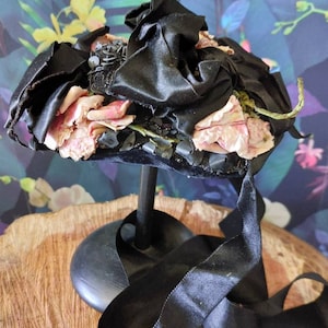 Pu&ograve; includere: Un cappello di velluto nero con rose di tessuto rosa e perline nere. Il cappello &egrave; su un supporto nero con un nastro nero.