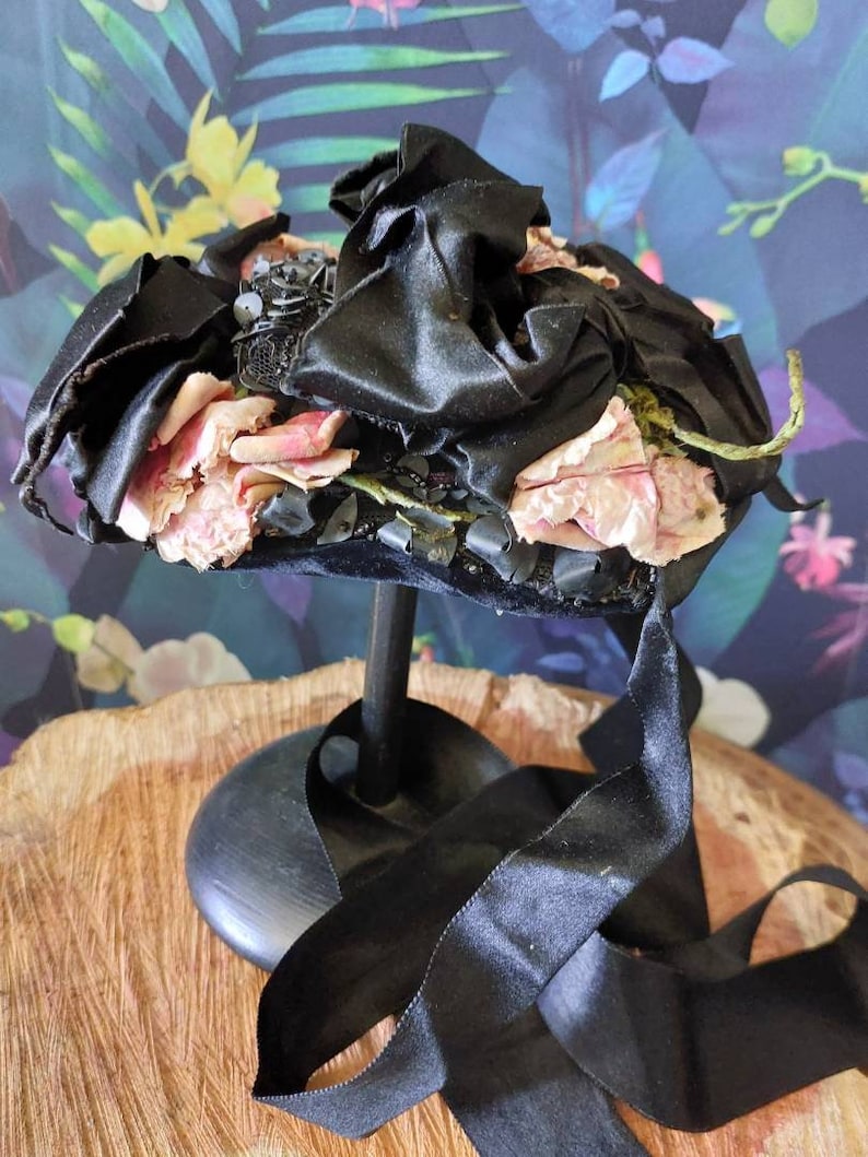 Pu&ograve; includere: Un cappello di velluto nero con rose di tessuto rosa e perline nere. Il cappello &egrave; su un supporto nero con un nastro nero.