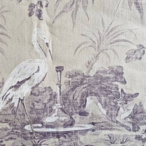 May include: Fabric with a toile design depicting a white heron, a decorative bottle, and foliage in shades of purple and grey. The background is a light beige colour, creating a vintage aesthetic.