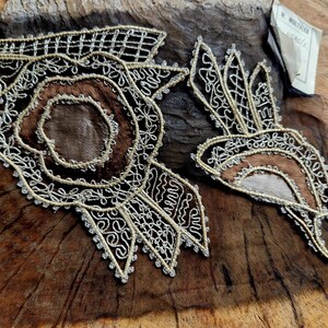 May include: Two intricate lace designs, one circular and one more free-flowing, are stitched onto a piece of brown leather. The designs are made with gold thread and have a delicate, almost floral appearance.