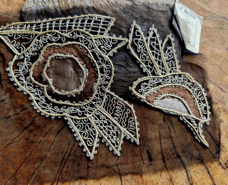May include: Two intricate lace designs, one circular and one more free-flowing, are stitched onto a piece of brown leather. The designs are made with gold thread and have a delicate, almost floral appearance.