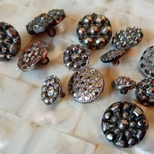 May include: A collection of 13 silver-toned buttons with various embossed designs. The buttons are round and have a raised, textured surface.