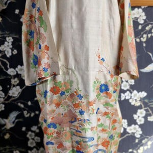May include: A vintage kimono in cream with a colorful floral and landscape design. The long sleeves and hem are adorned with blue, orange, and green flowers, trees, and bridges. The kimono is displayed on a mannequin.