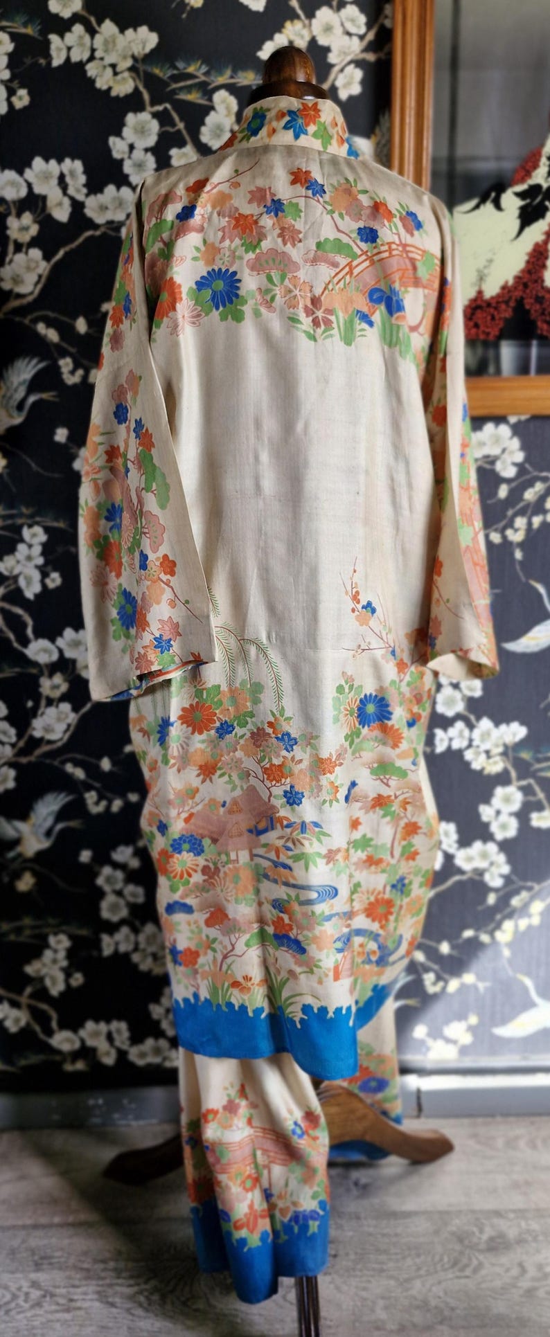 May include: A vintage kimono in cream with a colorful floral and landscape design. The long sleeves and hem are adorned with blue, orange, and green flowers, trees, and bridges. The kimono is displayed on a mannequin.