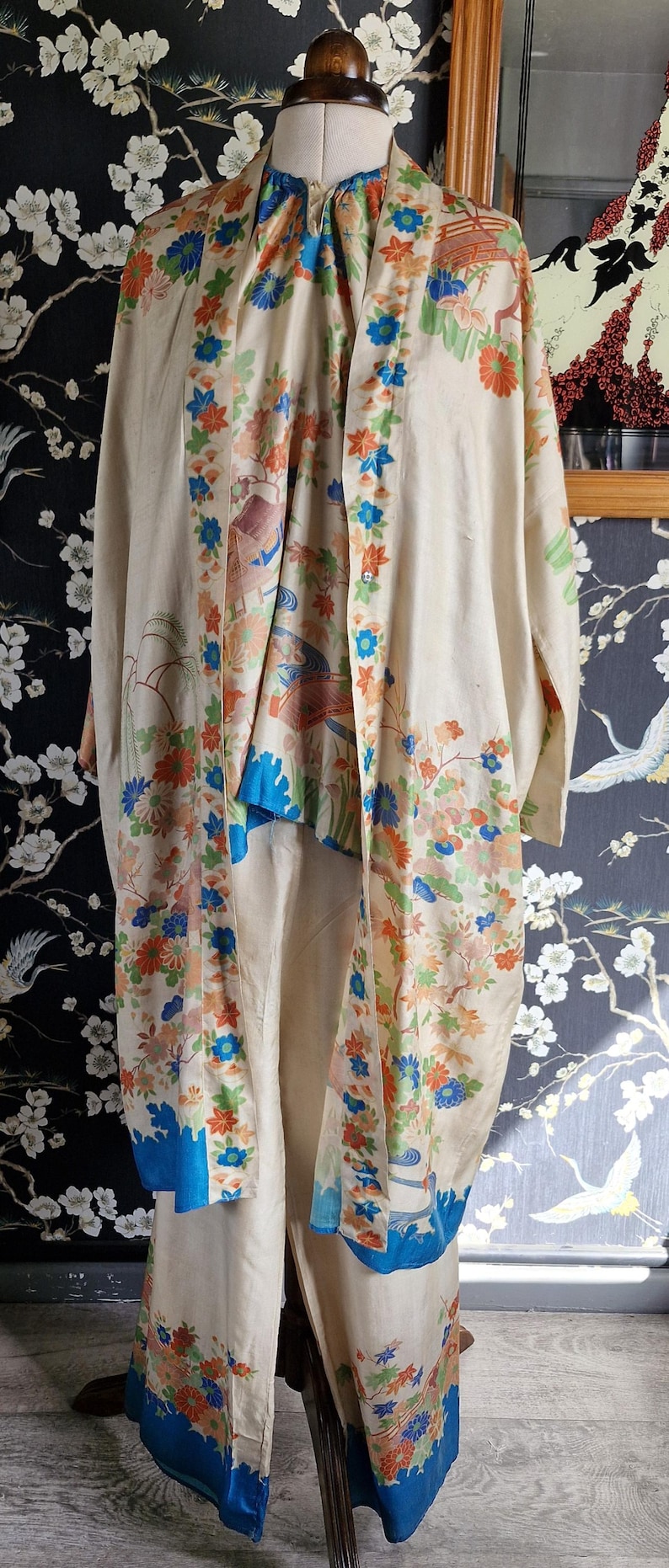 May include: Cream-colored vintage kimono set with a floral pattern in orange, blue, and green. The set includes a long robe and matching pants. The kimono is displayed on a mannequin.