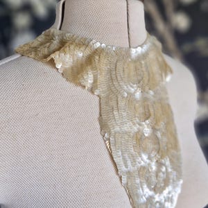 May include: A vintage-style, cream-colored sequin collar. The collar is displayed on a mannequin, showcasing its intricate design and shimmering texture. The sequins are arranged in a layered pattern, creating a cascading effect.
