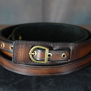 Peut inclure: Une ceinture en cuir marron avec une boucle en laiton et une sangle en cuir marron. La ceinture est enroulée et présente un motif cousu.
