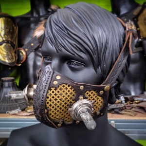 May include: A steampunk-style mask made of brown leather, brass, and metal components. The mask features a decorative brass mesh with a cross pattern and is secured with leather straps. The mask is displayed on a black mannequin head.