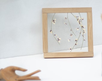 以下が含まれることがあります： 正方形の木製フレームのアートワーク。白い花と紫の花のデザインです。白い表面に置かれ、背景にはディフューザーと木の棒があります。