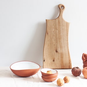 Op de afbeelding: Een houten snijplank met een unieke vorm en een handvat, een terracotta kom met een witte binnenkant, een stapel terracotta kommen, twee granaatappels en twee citroenen op een wit tafelkleed.
