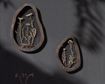 May include: Two wooden wall decor pieces in the shape of an irregular oval, each with a pressed dried flower arrangement inside. The pieces are mounted on a dark gray wall.