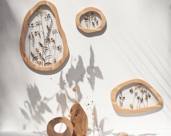 May include: Three wooden frames with irregular shapes, each containing pressed dried flowers and leaves. The frames are hanging on a white wall. There are two small wooden vases and a few stones on a white surface below the frames.