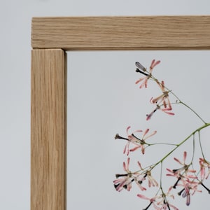 Pode incluir: Uma moldura de madeira com um design de flores prensadas. As flores s&atilde;o rosa e brancas com caules escuros. A moldura &eacute; feita de madeira marrom claro.