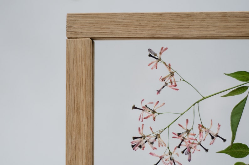 Pode incluir: Uma moldura de madeira com um design de flores prensadas. As flores s&atilde;o rosa e brancas com caules escuros. A moldura &eacute; feita de madeira marrom claro.