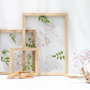 Pode incluir: Tr&ecirc;s molduras de madeira de tamanhos diferentes, cada uma com um arranjo de flores prensadas. As molduras est&atilde;o dispostas em forma de escada sobre uma superf&iacute;cie branca.