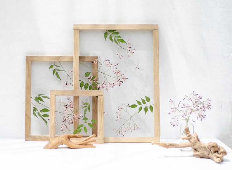 Pode incluir: Tr&ecirc;s molduras de madeira de tamanhos diferentes, cada uma com um arranjo de flores prensadas. As molduras est&atilde;o dispostas em forma de escada sobre uma superf&iacute;cie branca.