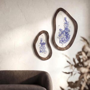 May include: Two wooden framed wall art pieces with an organic shape. Each frame holds a delicate arrangement of blue and white flowers. The frames are a warm brown color, and the background is a neutral white.