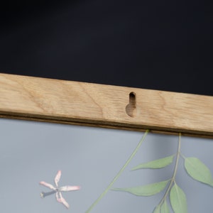 Pode incluir: Uma moldura de madeira com acabamento marrom claro. A moldura tem um furo para pendurar na parte superior e uma frente de vidro transparente. A moldura est&aacute; vazia, mas h&aacute; uma pequena flor seca e um ramo de folhas verdes dentro.