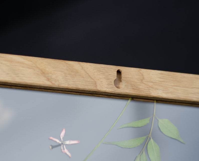 Pode incluir: Uma moldura de madeira com acabamento marrom claro. A moldura tem um furo para pendurar na parte superior e uma frente de vidro transparente. A moldura est&aacute; vazia, mas h&aacute; uma pequena flor seca e um ramo de folhas verdes dentro.