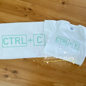 Puede incluir: Dos camisetas blancas con texto verde. La camiseta más grande dice "CTRL + C" y la más pequeña dice "CTRL + V".