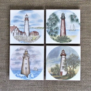May include: Set of four ceramic tile coasters featuring watercolor paintings of lighthouses. Each coaster depicts a different lighthouse with a unique design and color scheme.
